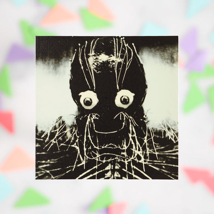 A square black and white art print featuring a creepy spider-like face with large white eyes and web-like strands, centered on a white marble background with blurred pastel paper confetti.